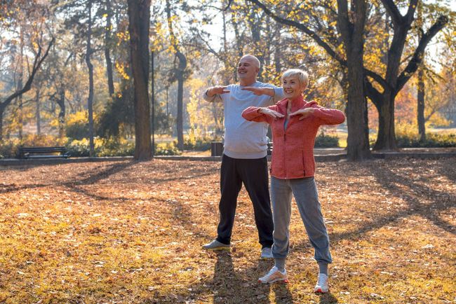 Two people doing arm exercises in a sunlit park with autumn trees and fallen leaves.