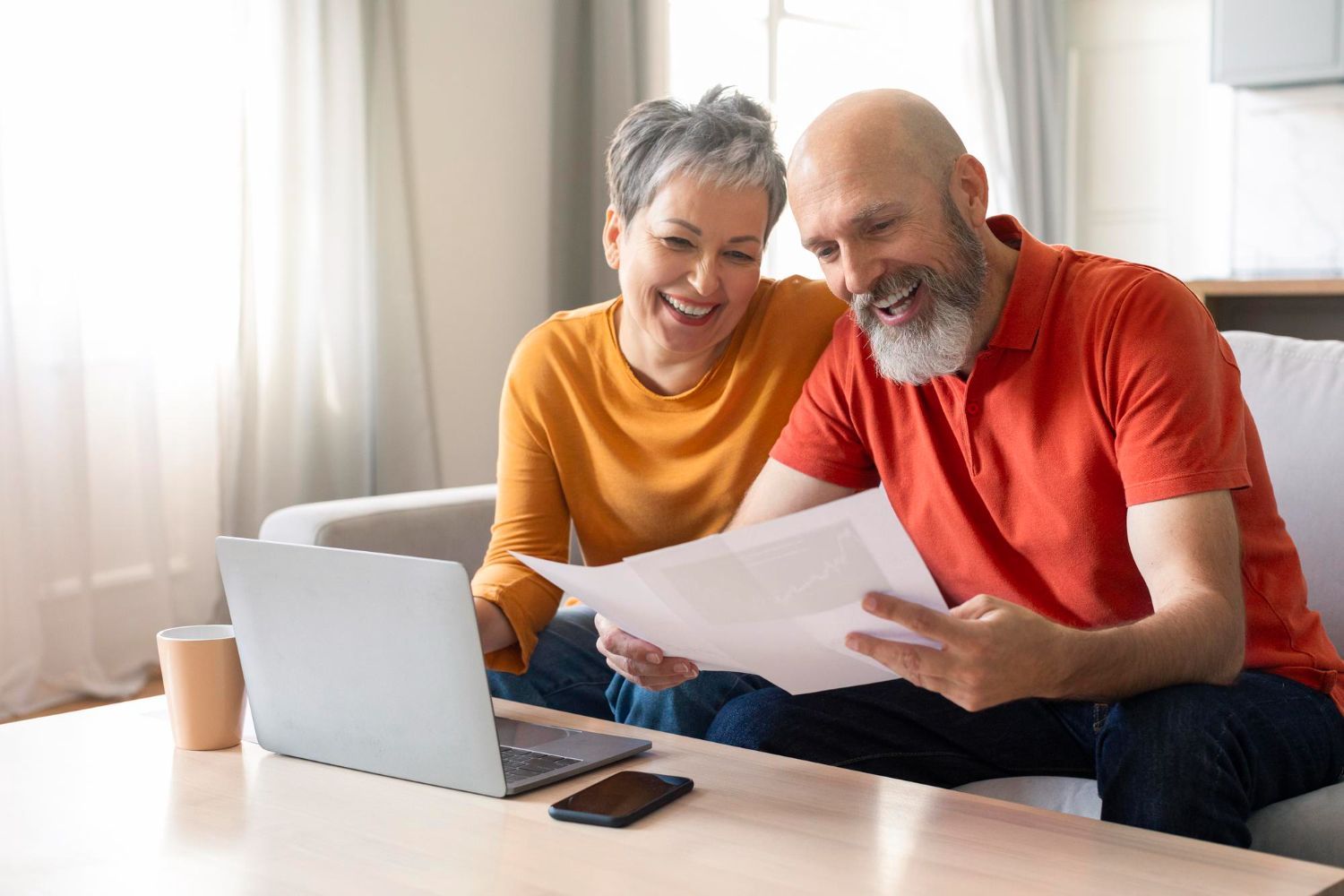 Two people smiling at a table, reading papers beside an open laptop and phone in a bright living room.