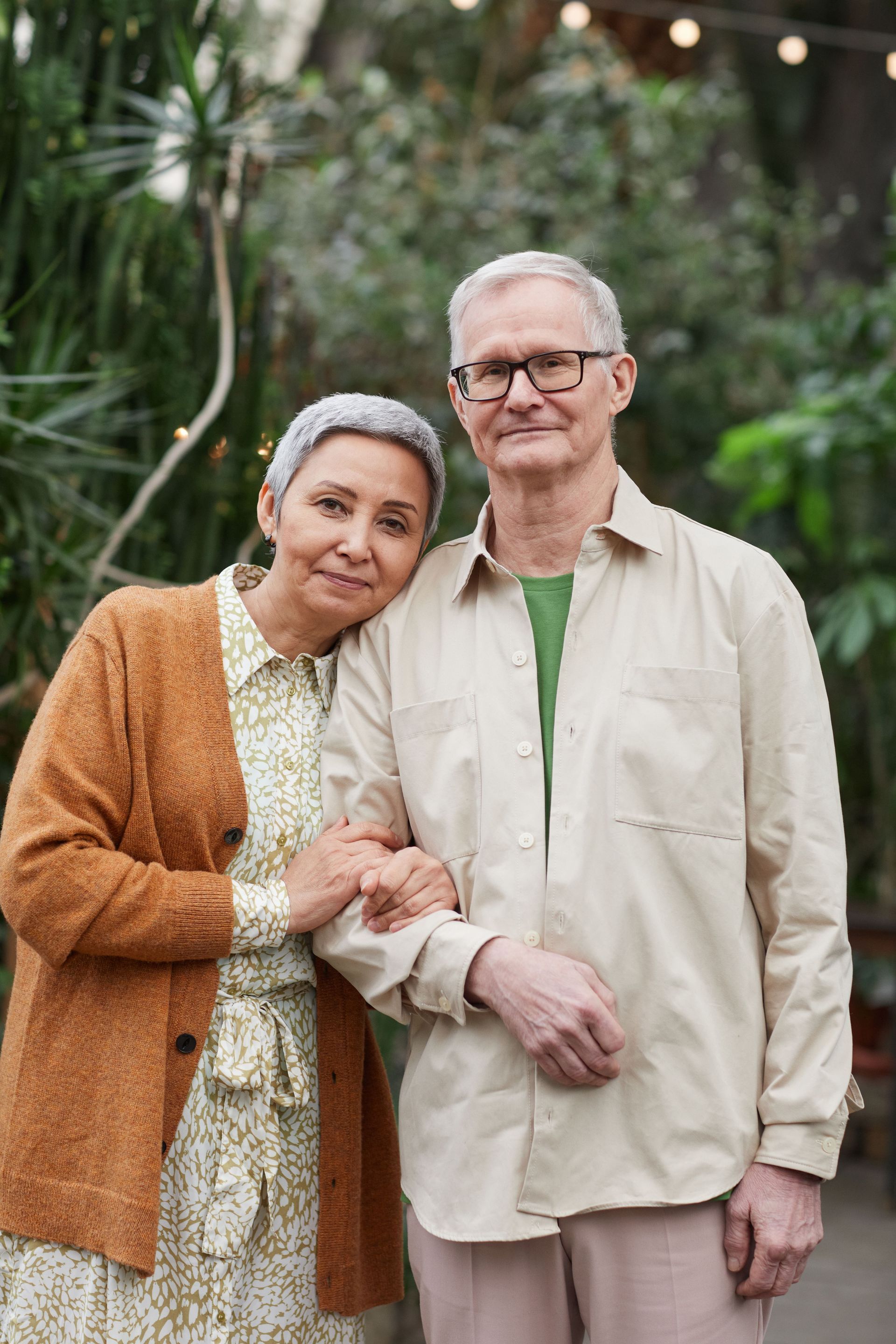 Two adults standing arm in arm in a garden, with soft string lights and greenery behind them.
