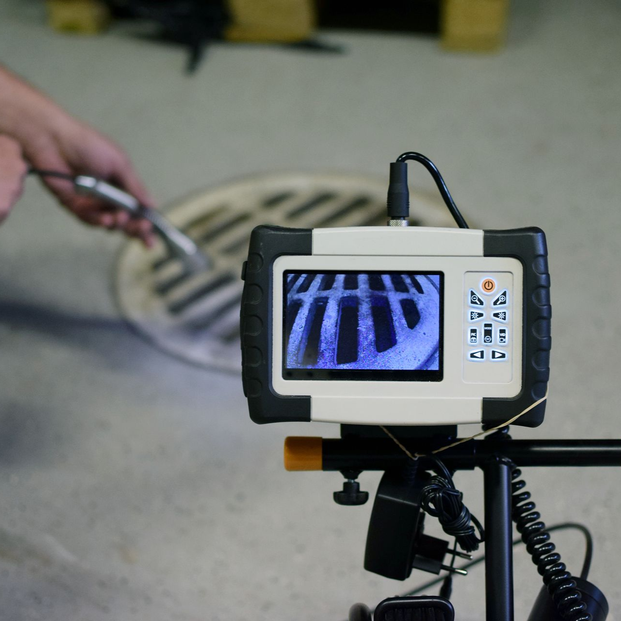 A close-up shows a screen displaying an inspection camera's view of a grate, held by a person in a concrete setting.