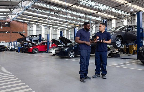 Two mechanics in blue uniforms converse in a car repair shop. Cars are on lifts.