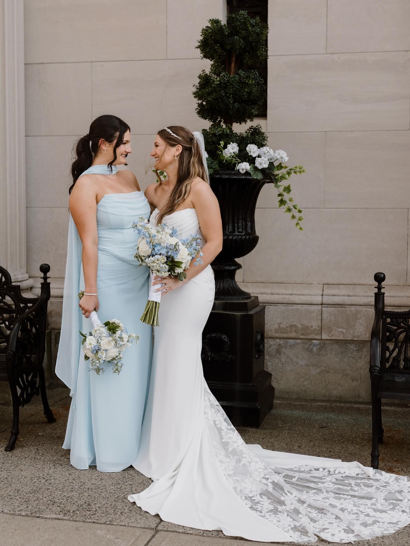 Two brides in pastel blue and white wedding dresses hold bouquets beside a stone wall and planter