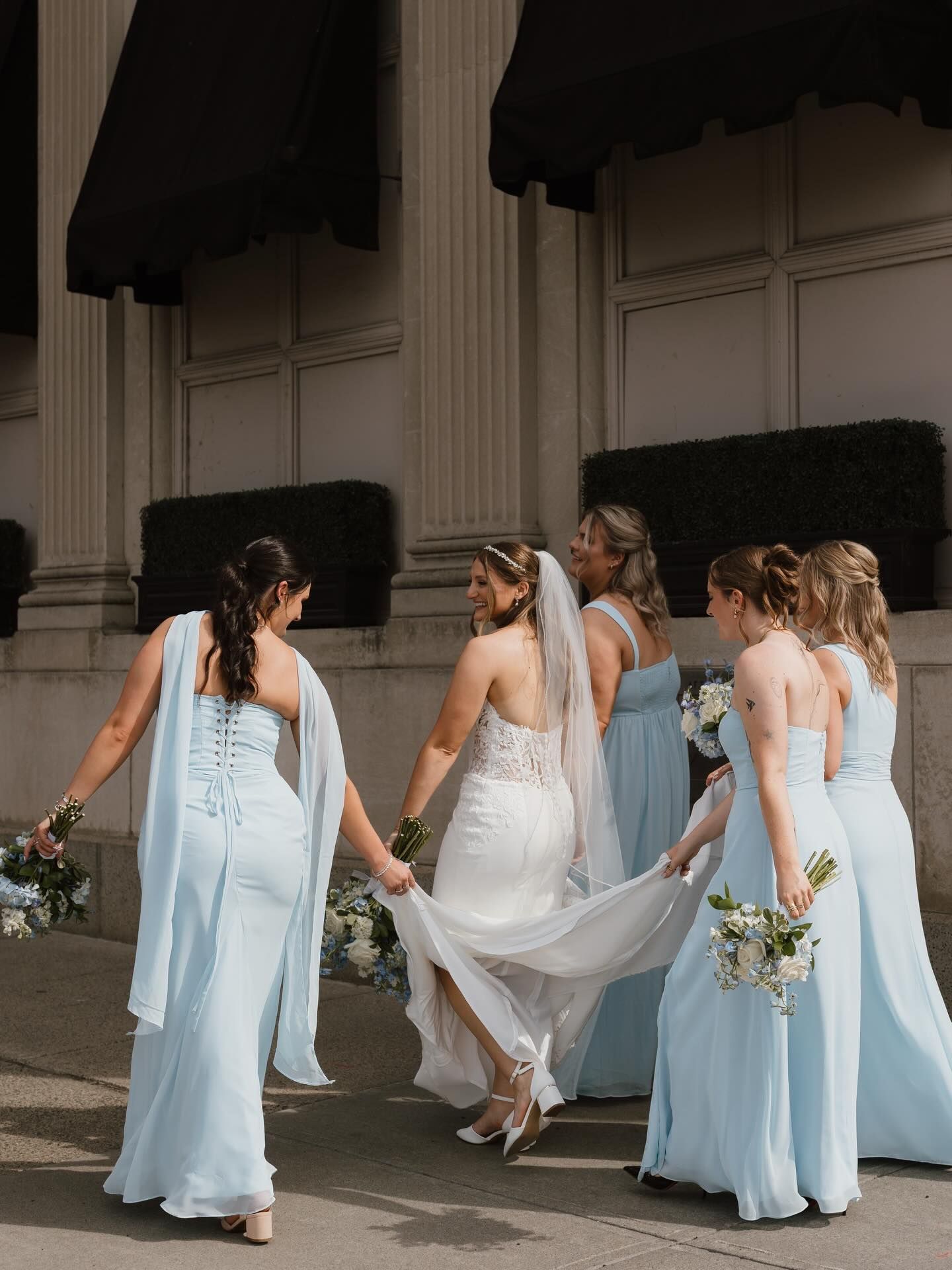 Five women in light blue dresses and white bridal gown walking together outdoors with bouquets