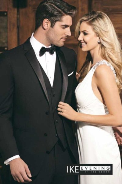 A groom in a black tuxedo and a bride in a white gown smiling at each other indoors.