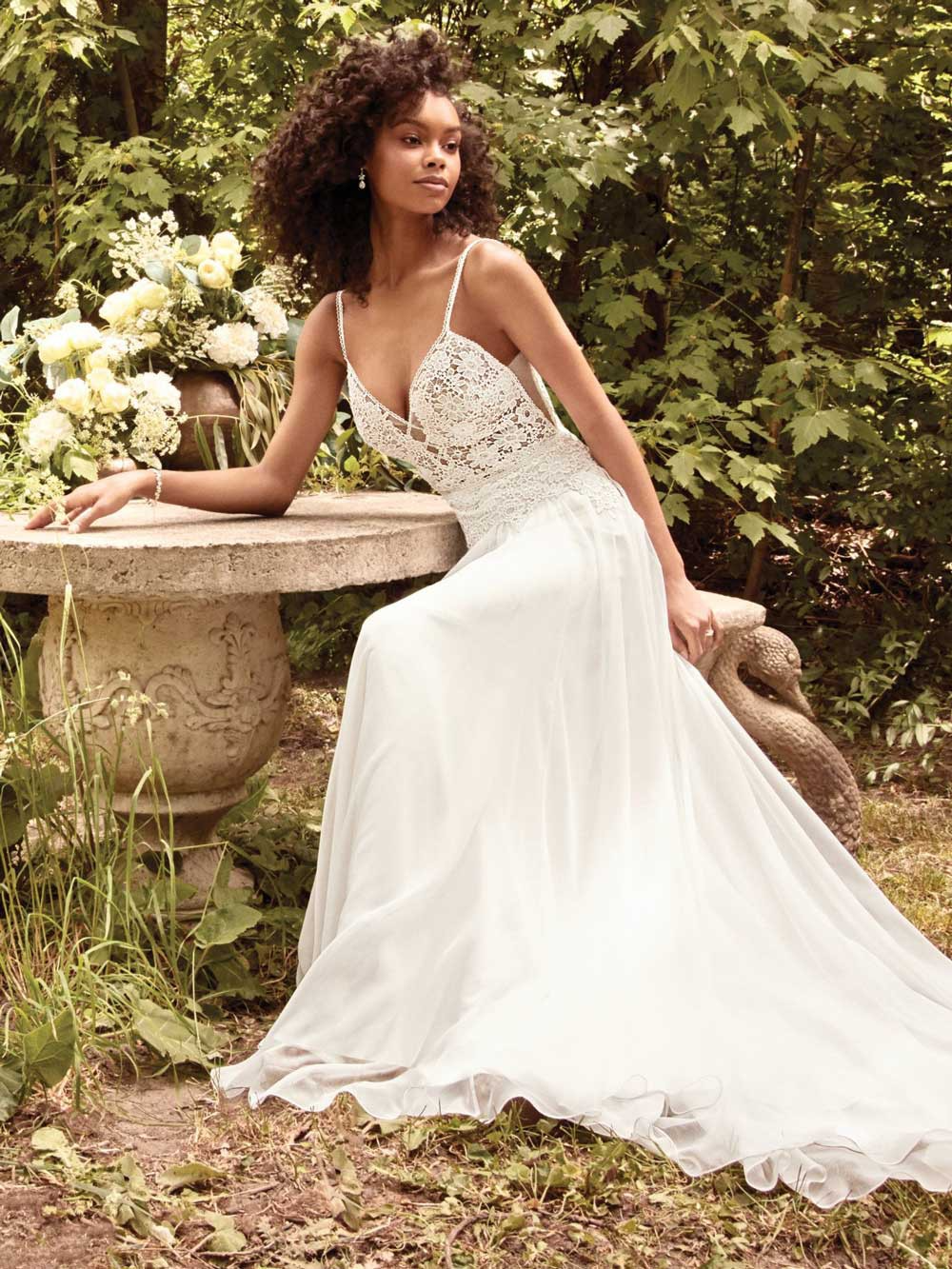 Woman in a white lace dress seated outdoors beside a stone table in a lush garden