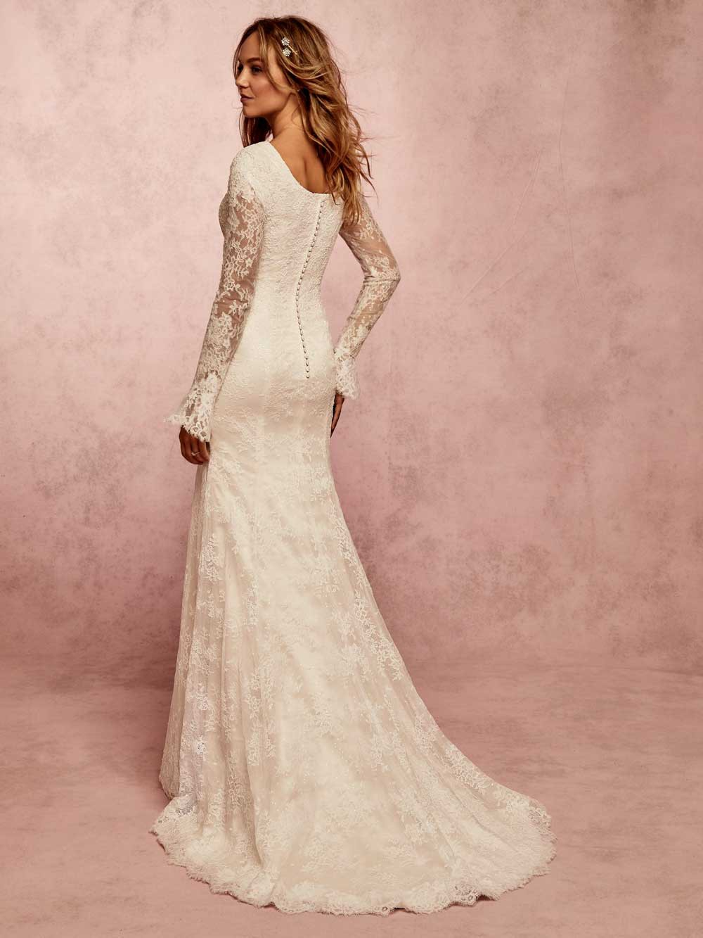 Woman in a long ivory lace gown standing in profile against a pink studio backdrop