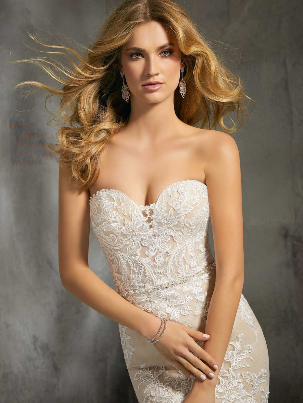 Woman in a strapless ivory lace gown against a gray backdrop, posing with flowing hair.