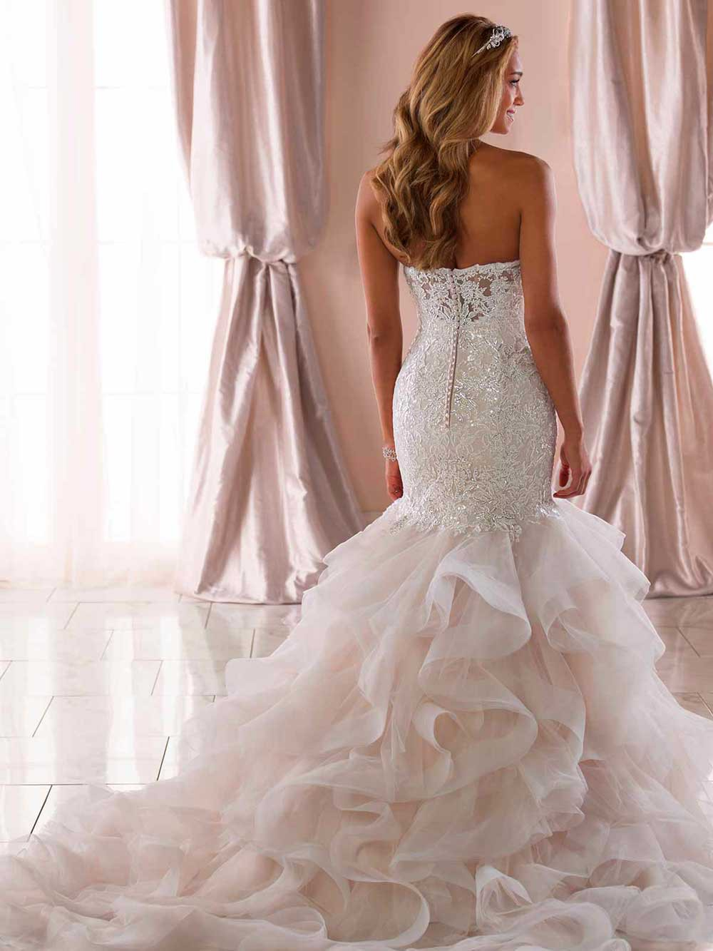 Woman in a white strapless wedding gown with a long ruffled train standing by sheer curtains