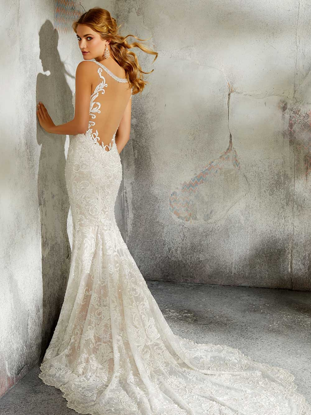 Bride in a white lace gown with a long train, standing by a textured wall.