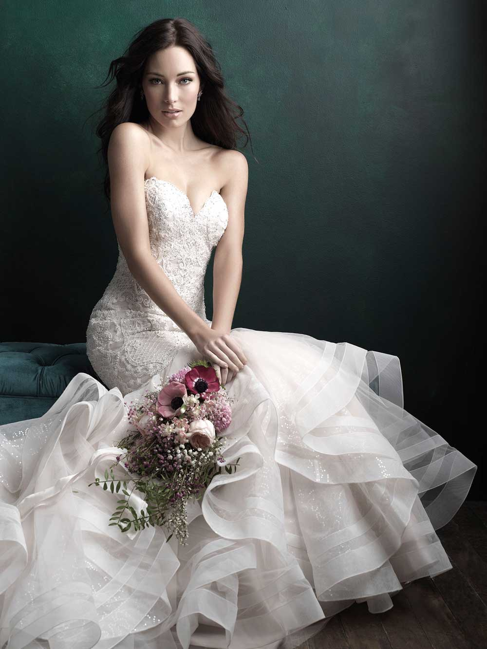 Woman in a white wedding dress holding a bouquet, seated against a dark backdrop.