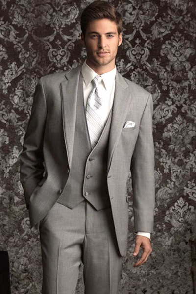 Man in a light gray three-piece suit standing against patterned wallpaper.