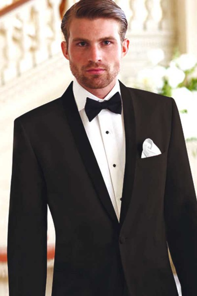 Man in a black tuxedo with bow tie and white pocket square, standing indoors on a staircase.