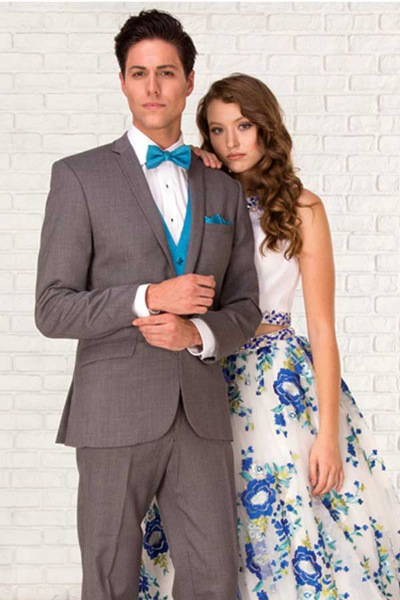 Two people posing against a white brick wall, wearing formal gray and floral attire.
