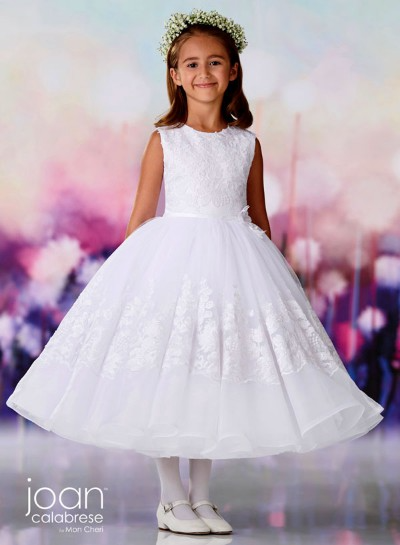 Girl in a white lace dress and floral headband, standing against a soft pastel backdrop.