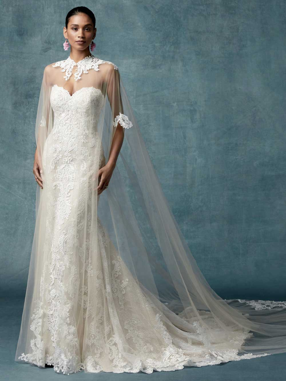 Woman in a white lace wedding dress with a long train and veil against a blue-gray studio backdrop