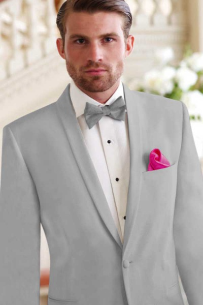 Man in a light gray tuxedo with a pink pocket square, standing indoors against a soft white staircase backdrop