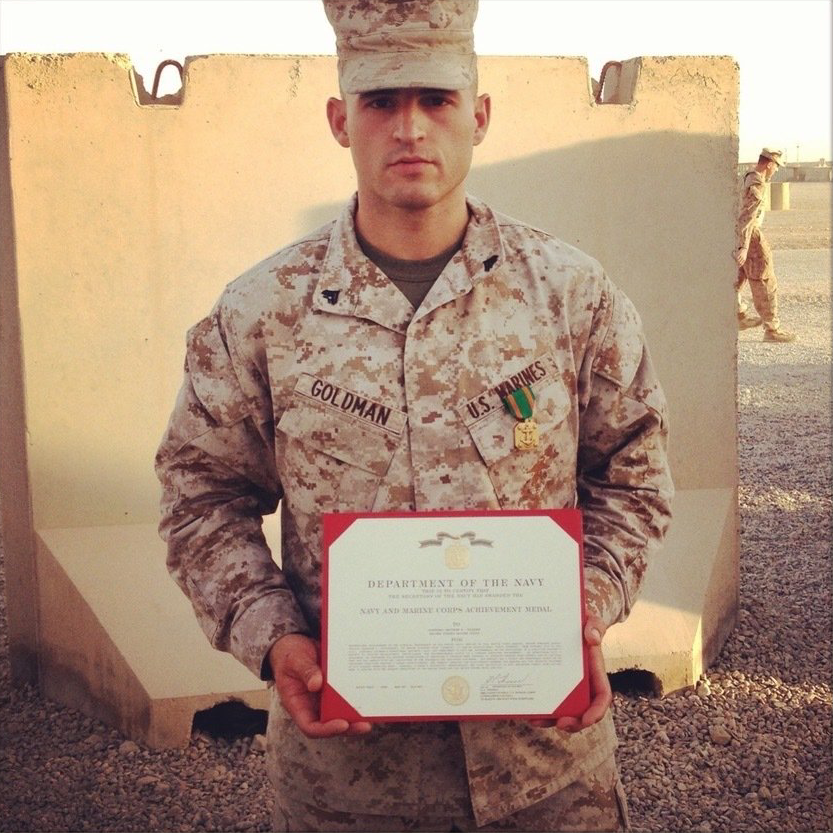 A service member in camouflage fatigues stands outdoors holding a certificate in front of a tan barrier.