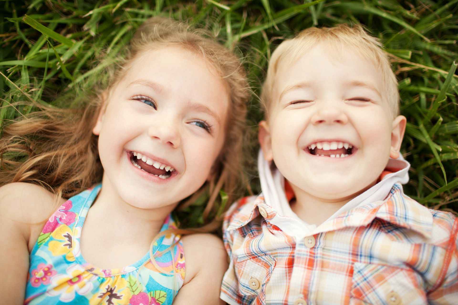 Due bambini sorridenti sdraiati sull’erba, uno con un vestito a fiori e l’altro con una camicia a quadri