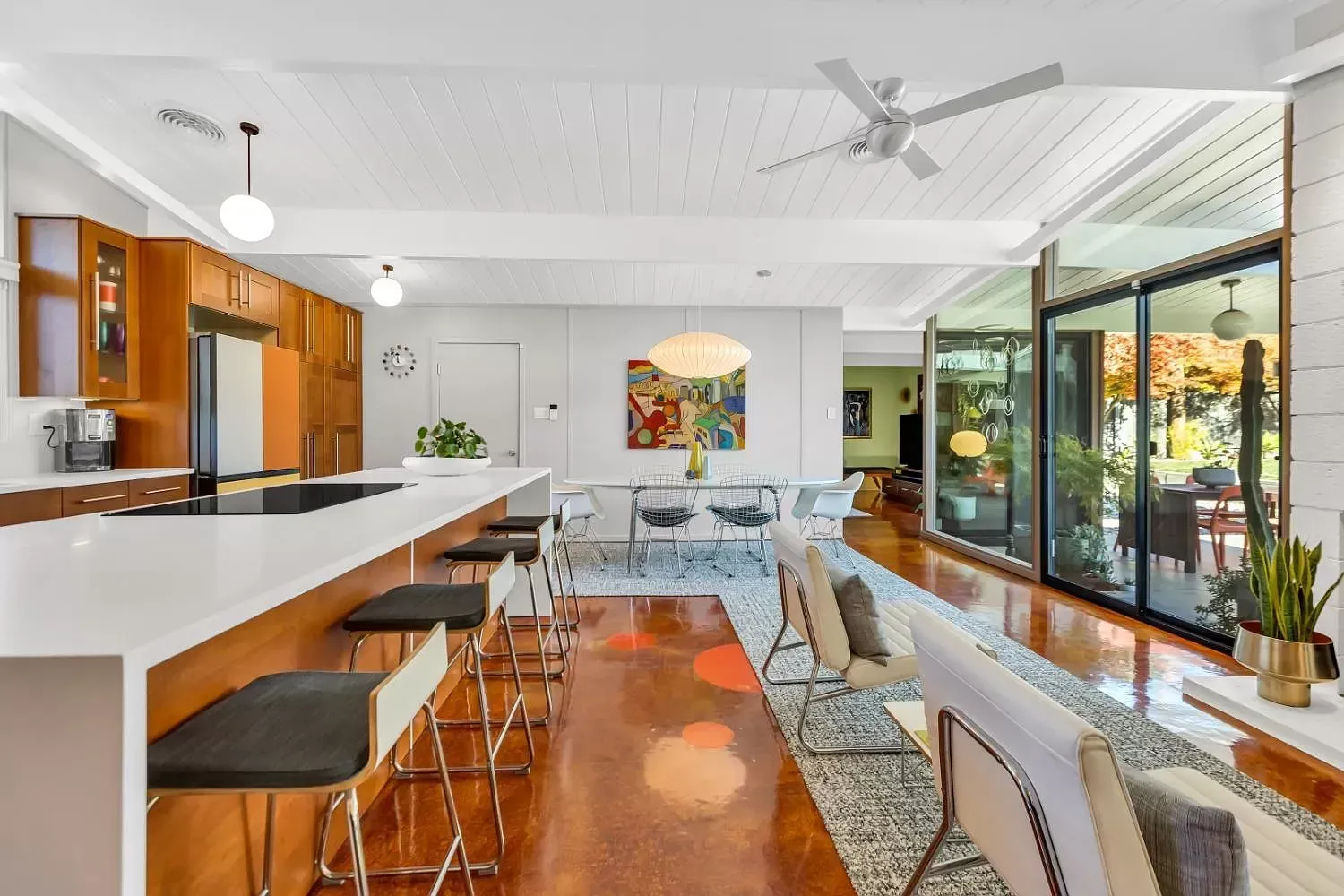 Bright open-plan living room and kitchen with white bar stools, wood floors, large windows, and indoor plants | Beth Ellinthorpe Realtor | Luxury Real Estate