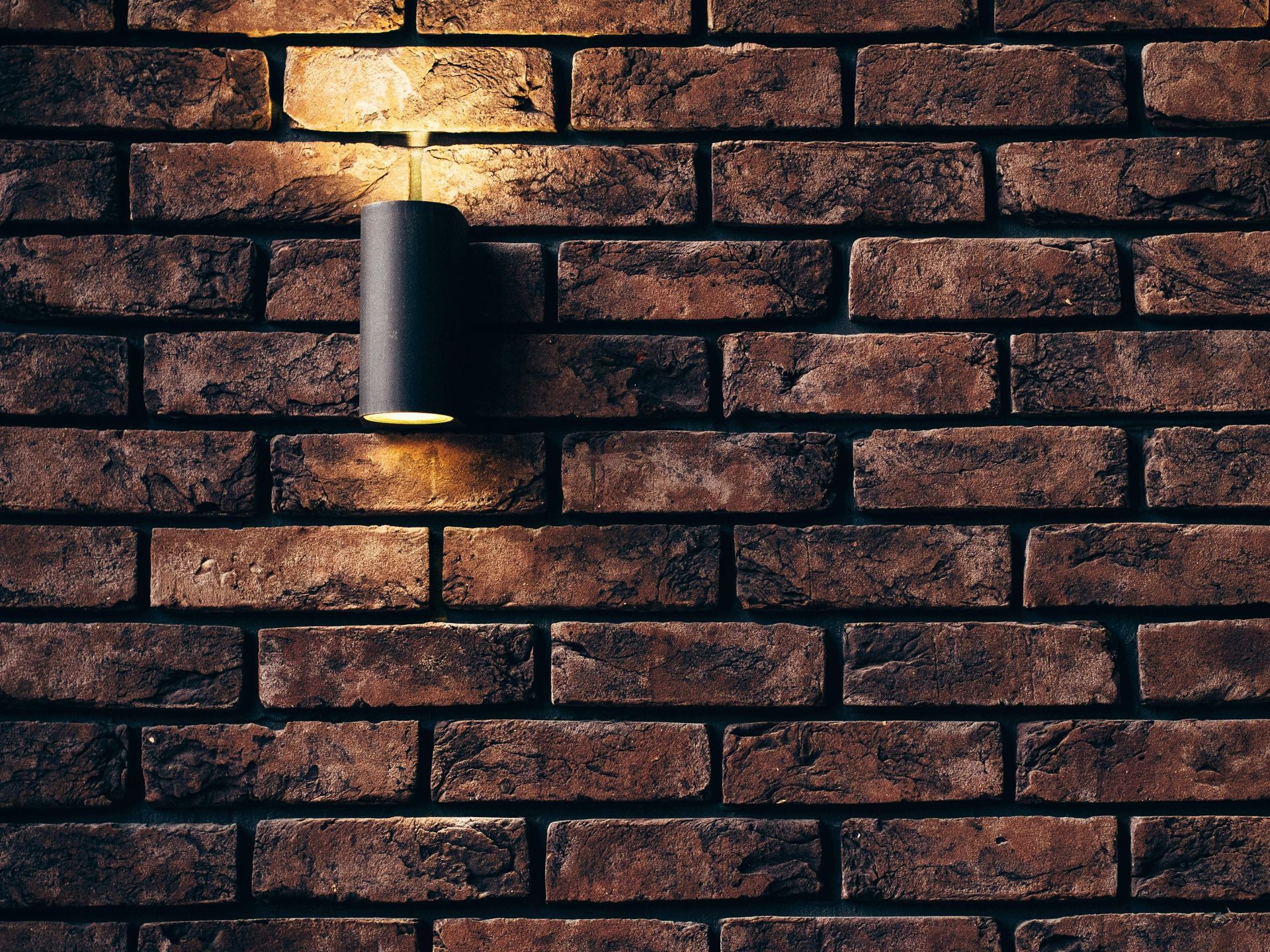 Black wall lamp on a weathered brick wall, illuminated with warm light.