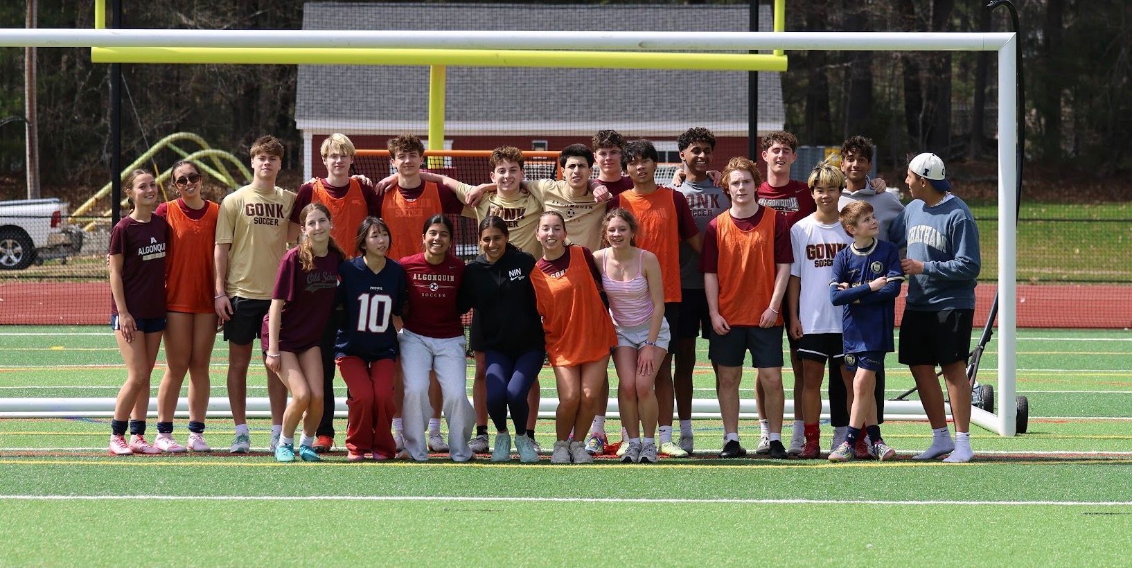Boys and girls lacrosse team photo posing on a field in front of a goal, wearing red, orange, and blue uniforms.