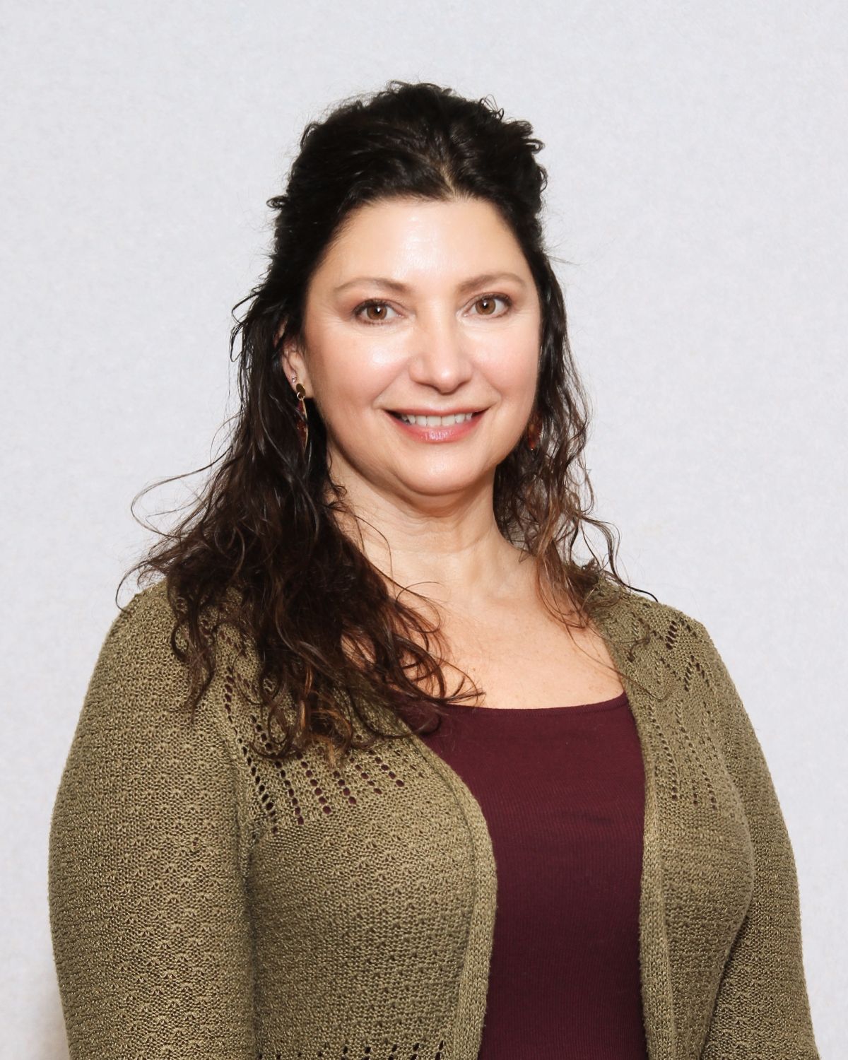 Woman with dark hair smiles, wearing a burgundy top and olive green cardigan, standing against a neutral background.