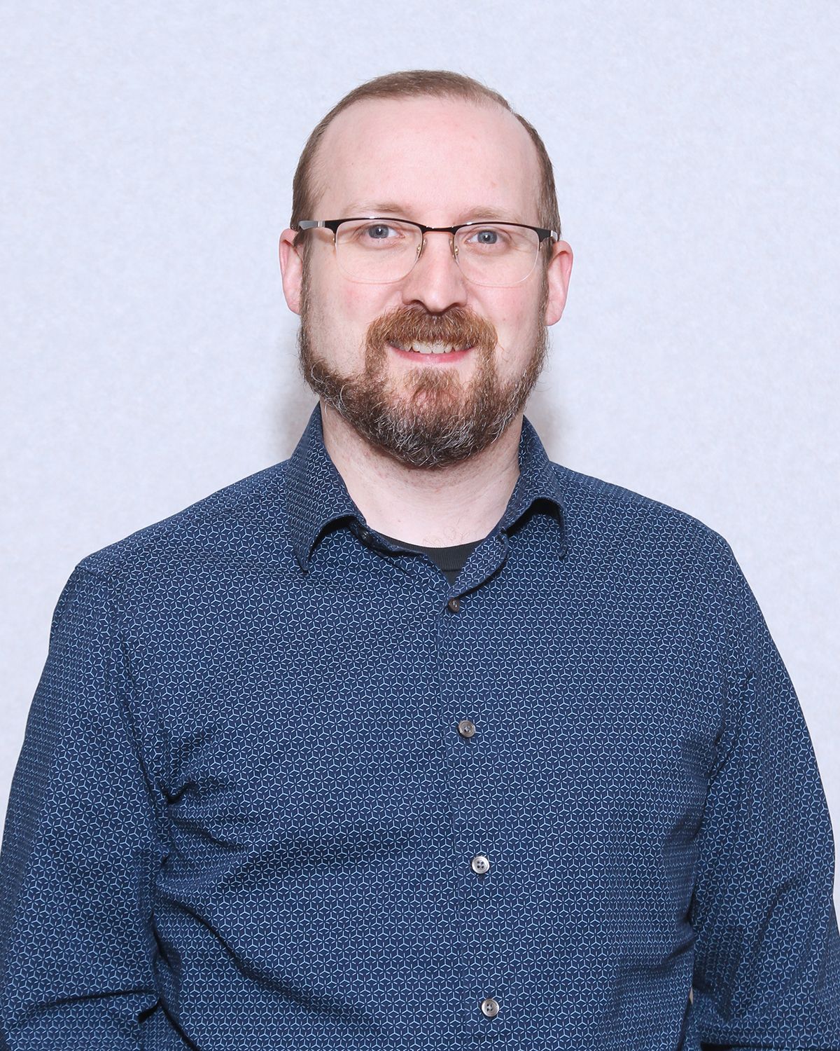Man with a beard, glasses, and blue patterned shirt smiles, looking at the camera against a white backdrop.