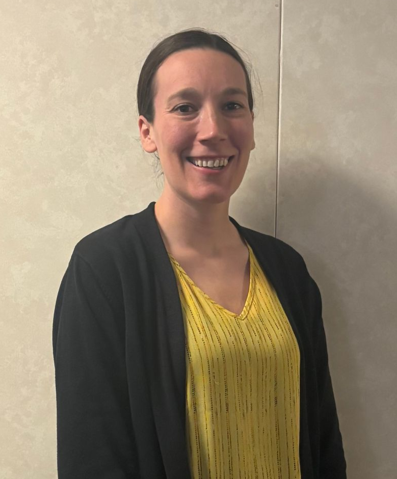 Woman in yellow top and black cardigan smiles, standing in front of a neutral-toned wall.