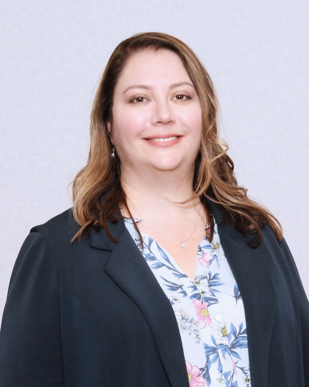 Woman smiling, wearing a blue blazer over a floral blouse, against a light gray backdrop.