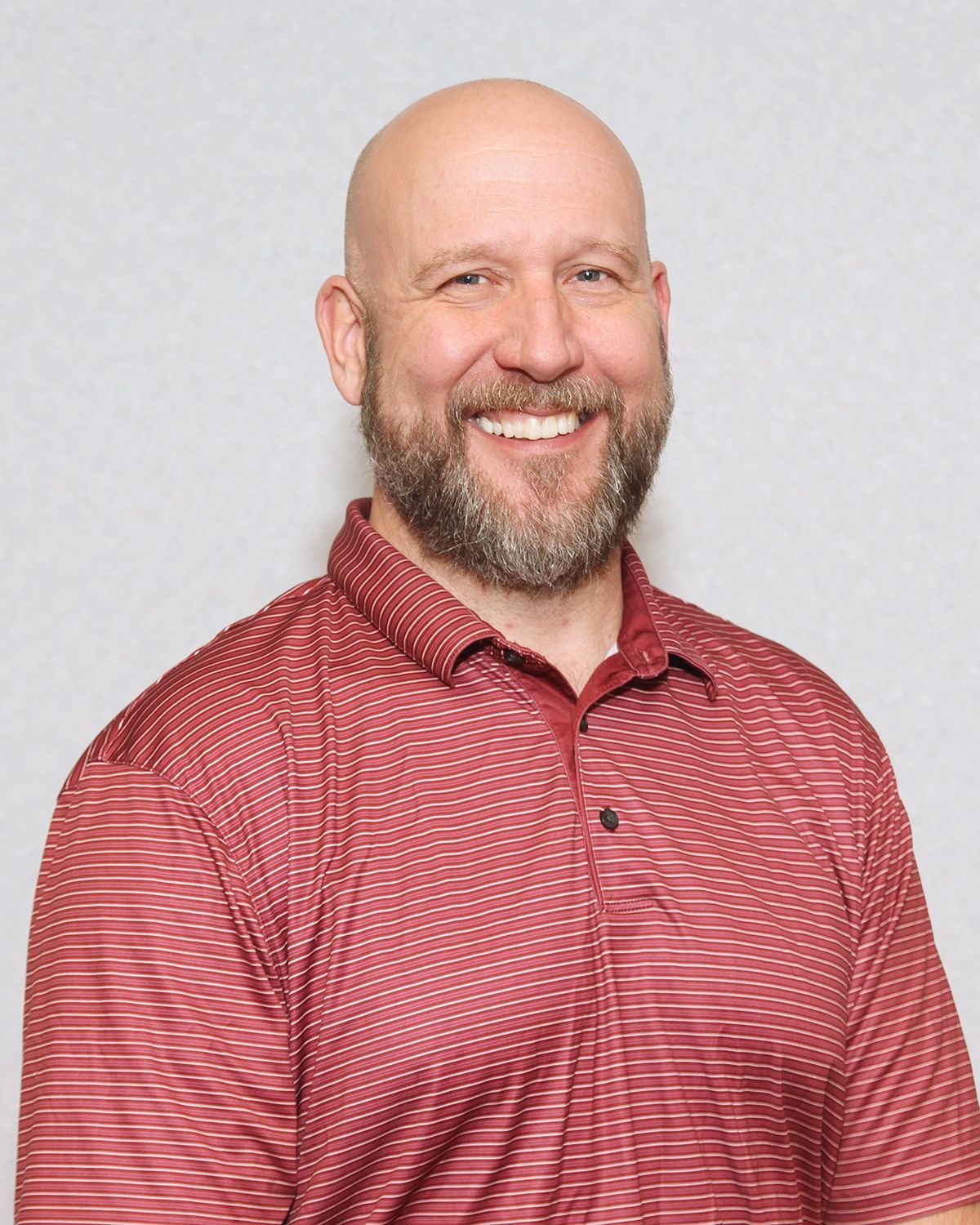 Bald man with beard smiles, wearing a red-striped polo shirt in front of a neutral backdrop.