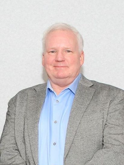 Man in gray blazer and blue shirt smiles, posing against a white backdrop.