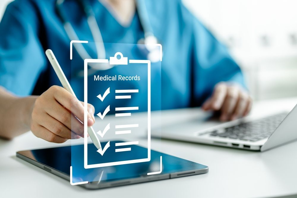 Hand checking medical record form on tablet beside laptop in a clinic setting
