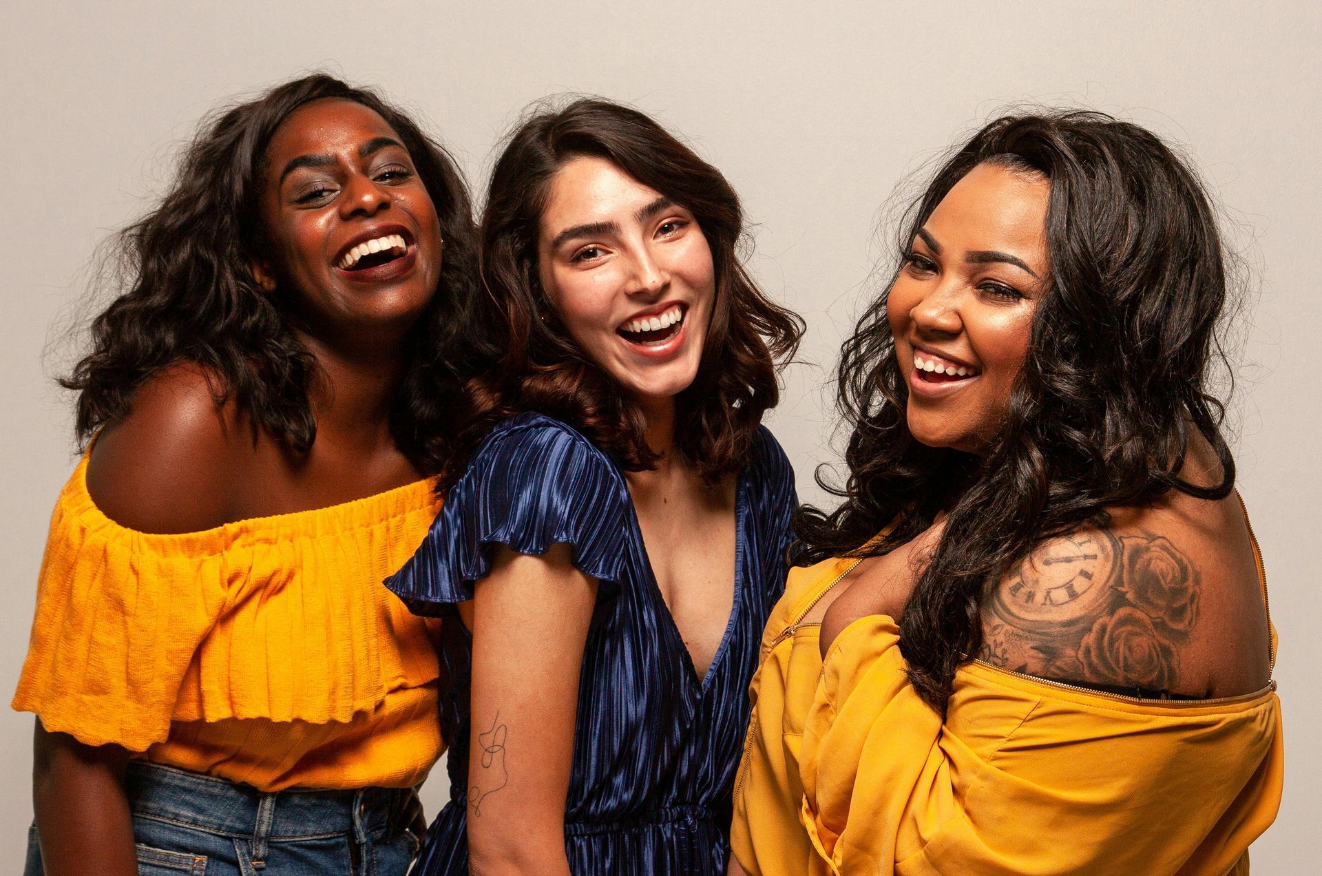 Three smiling women, one in blue dress, two in yellow tops. White background.