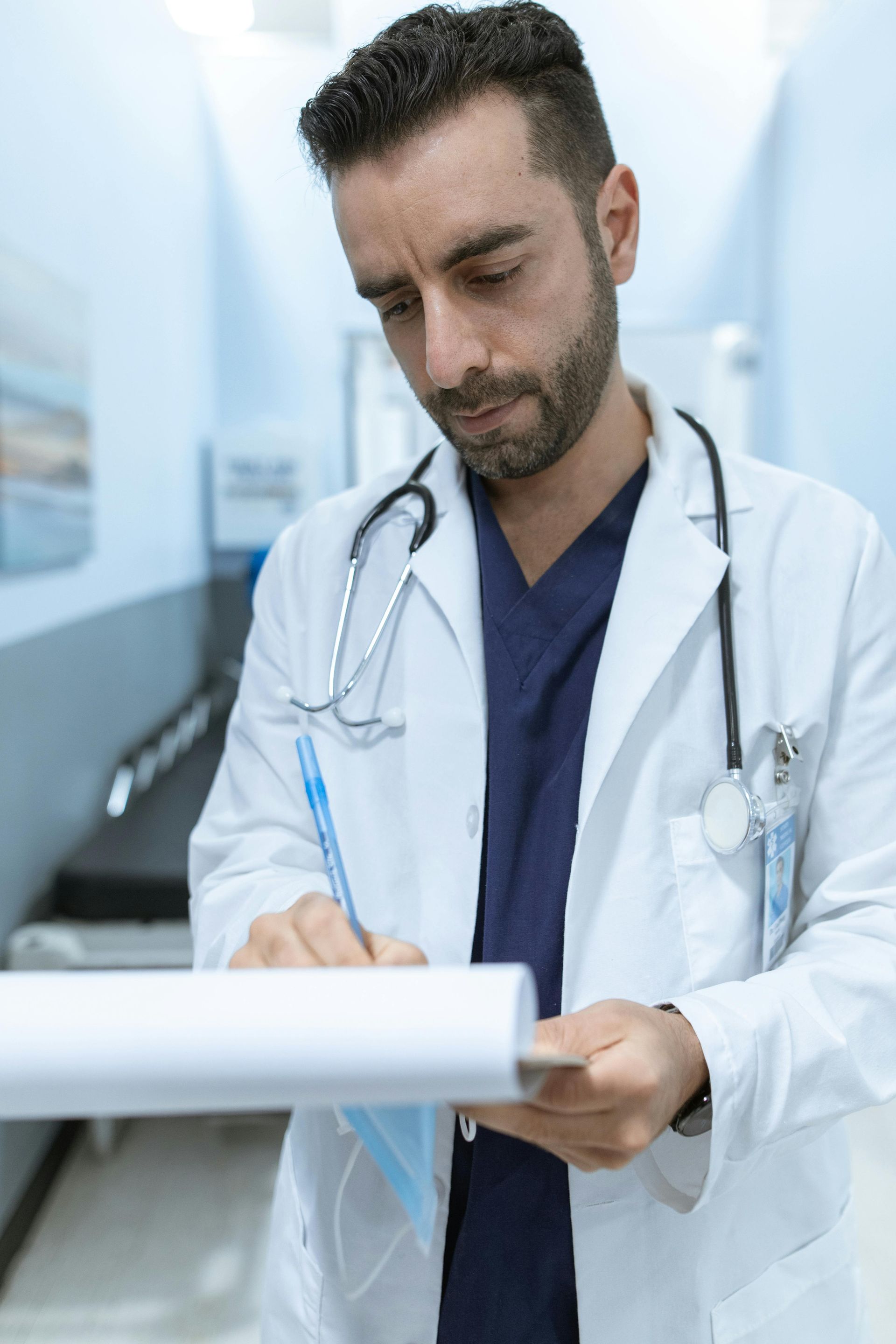 Doctor in white coat, writing on a clipboard in a hospital hallway.