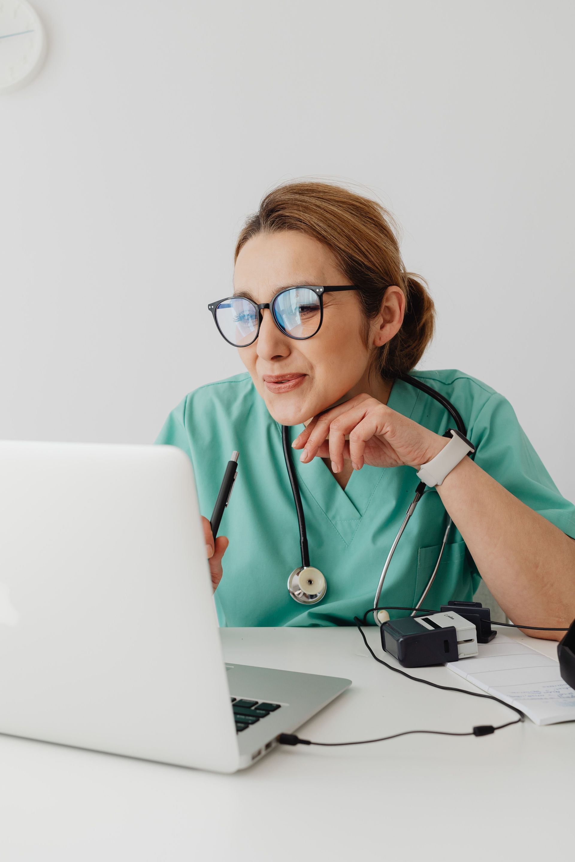 Healthcare worker in green scrubs and glasses using a laptop at a desk, with a stethoscope nearby