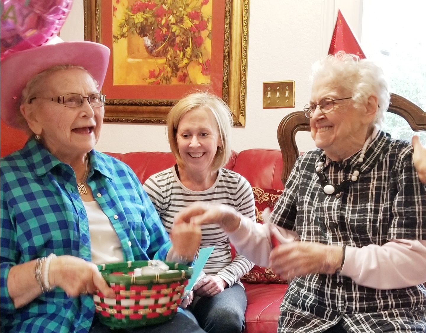 Two people wearing party hats sit on a red sofa, one holding a small basket, with a third person smiling between them.