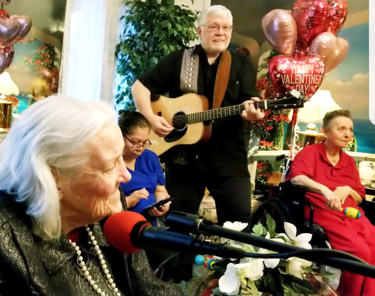 A guitarist performs for a group of people sitting indoors, with festive Valentine’s Day balloons in the background.