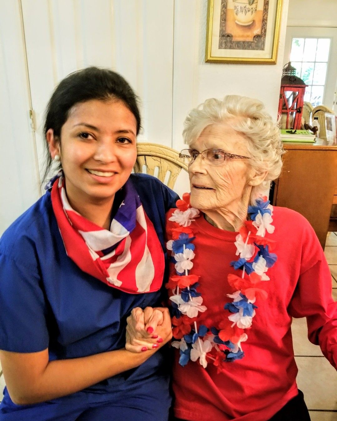 A person in blue scrubs with an American flag scarf holds hands with a person in a red shirt wearing a patriotic lei.