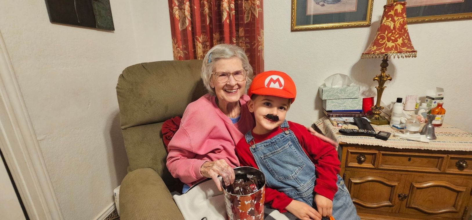 A person in a pink top and a child dressed as Mario sit in an armchair near a side table with a lamp.