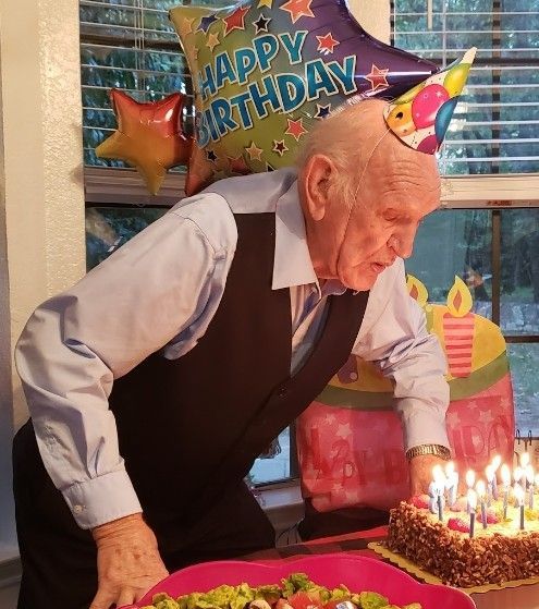 An elderly person in a party hat leans over a birthday cake with lit candles in a room decorated with balloons.