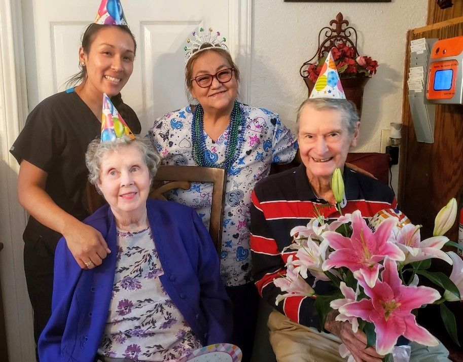 Four people wearing party hats, including one with a crown, smile while gathered for a celebration with a bouquet of lilies.