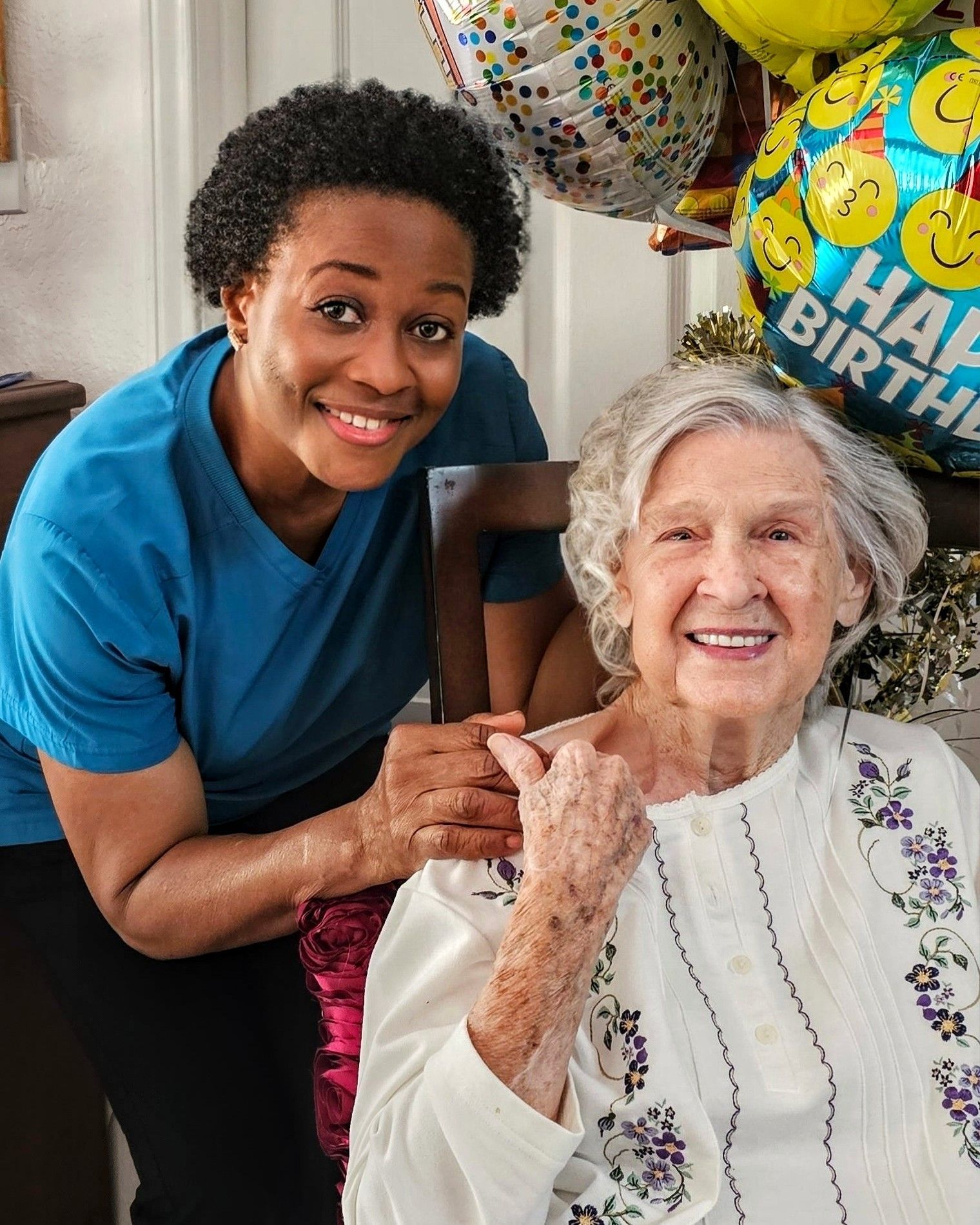 A smiling caregiver in blue scrubs holding the hand of an older person smiling in a white patterned top near balloons.