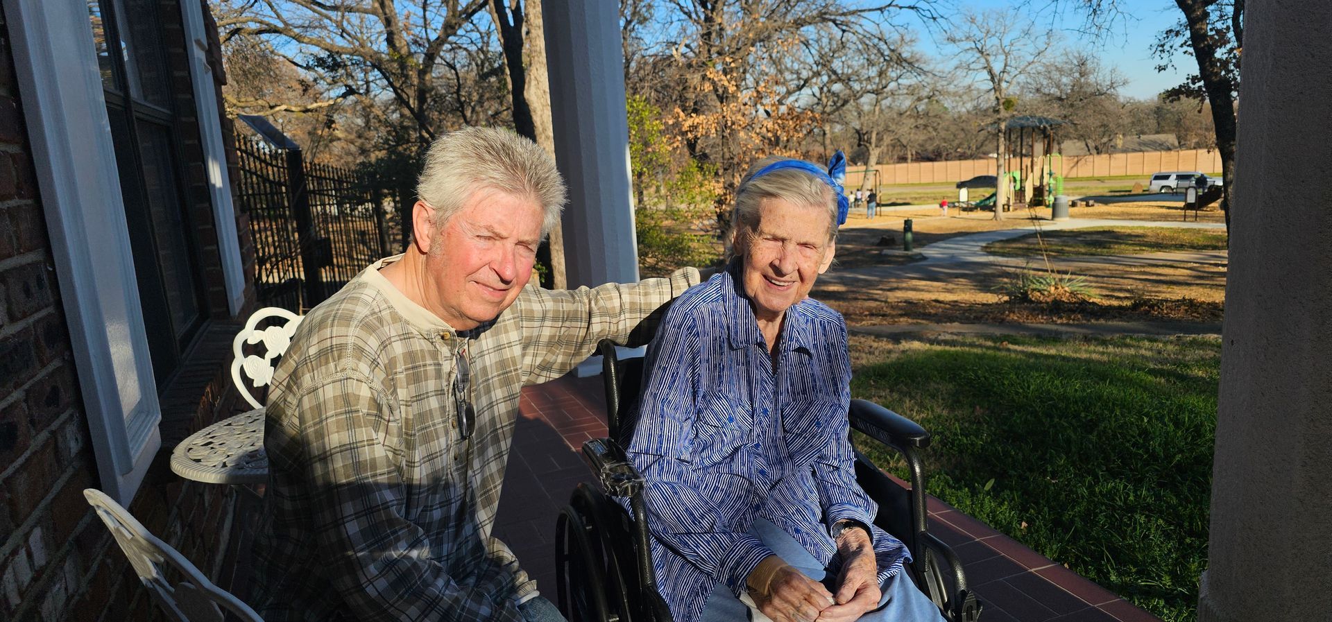 A person sitting in a wheelchair next to another individual on a sunlit porch overlooking a grassy landscape.