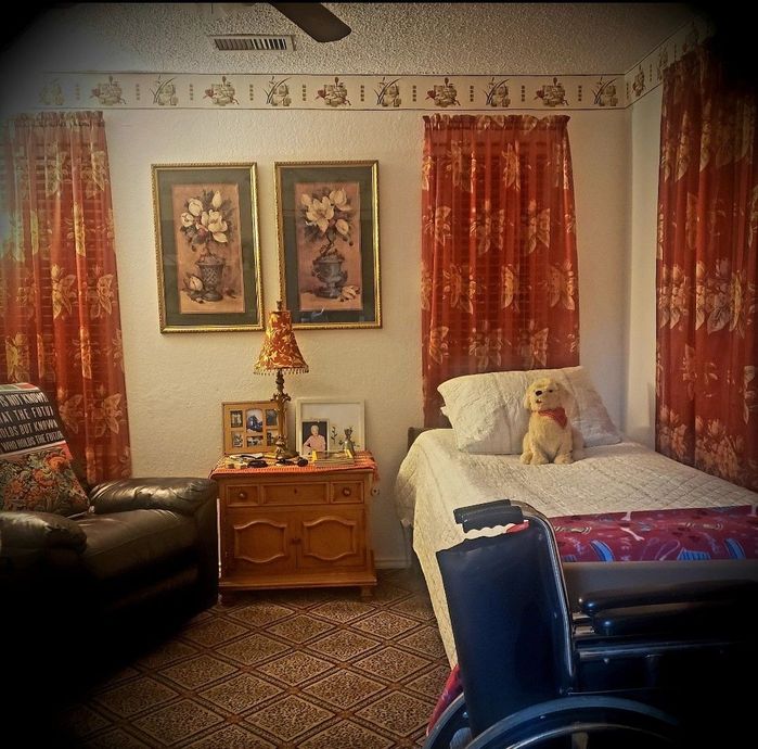 A room with a single bed, a recliner, a wooden nightstand, and a wheelchair, decorated with floral curtains and art.