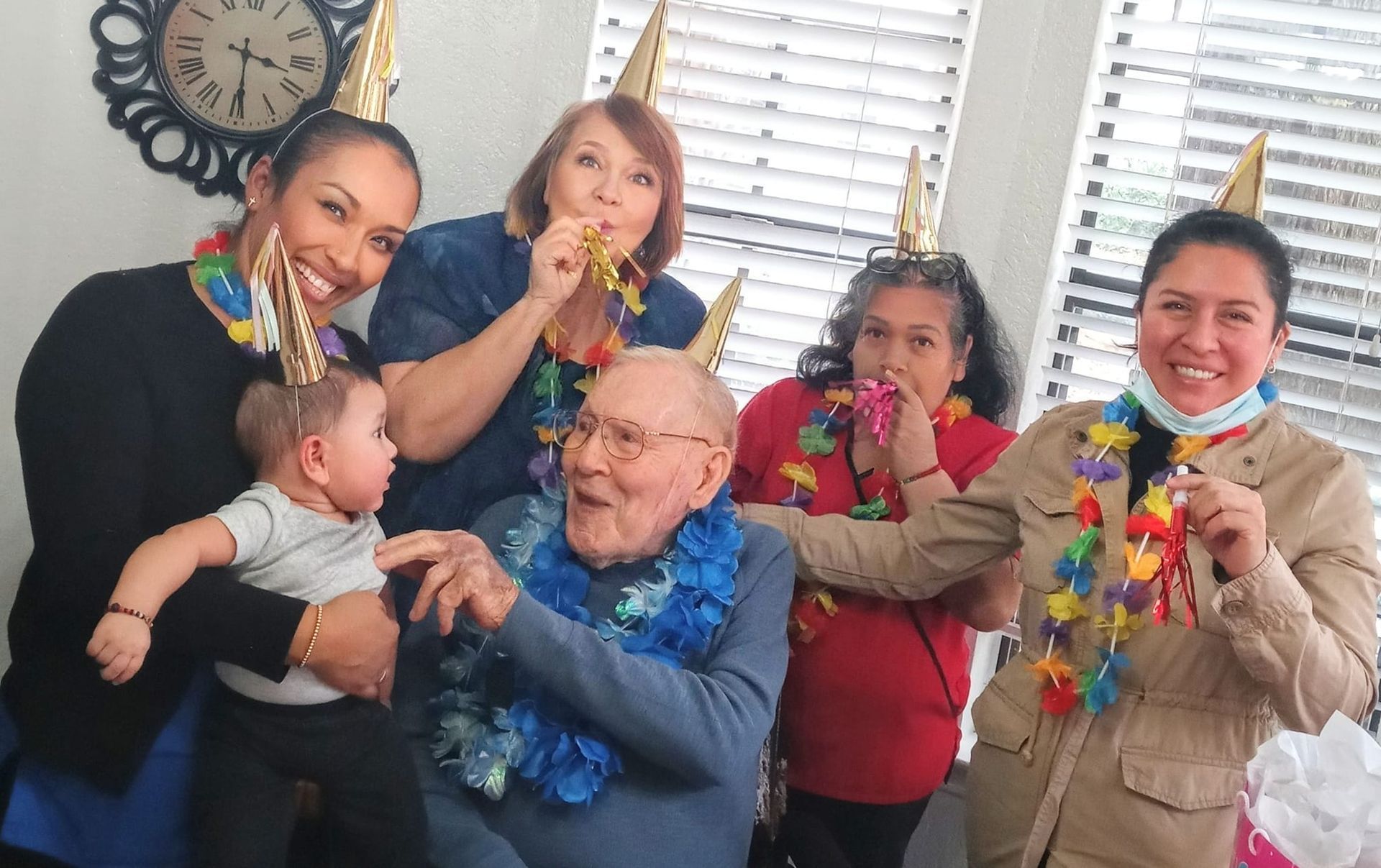 A group wearing party hats and leis celebrates around a seated person, with one holding a baby and others blowing horns.