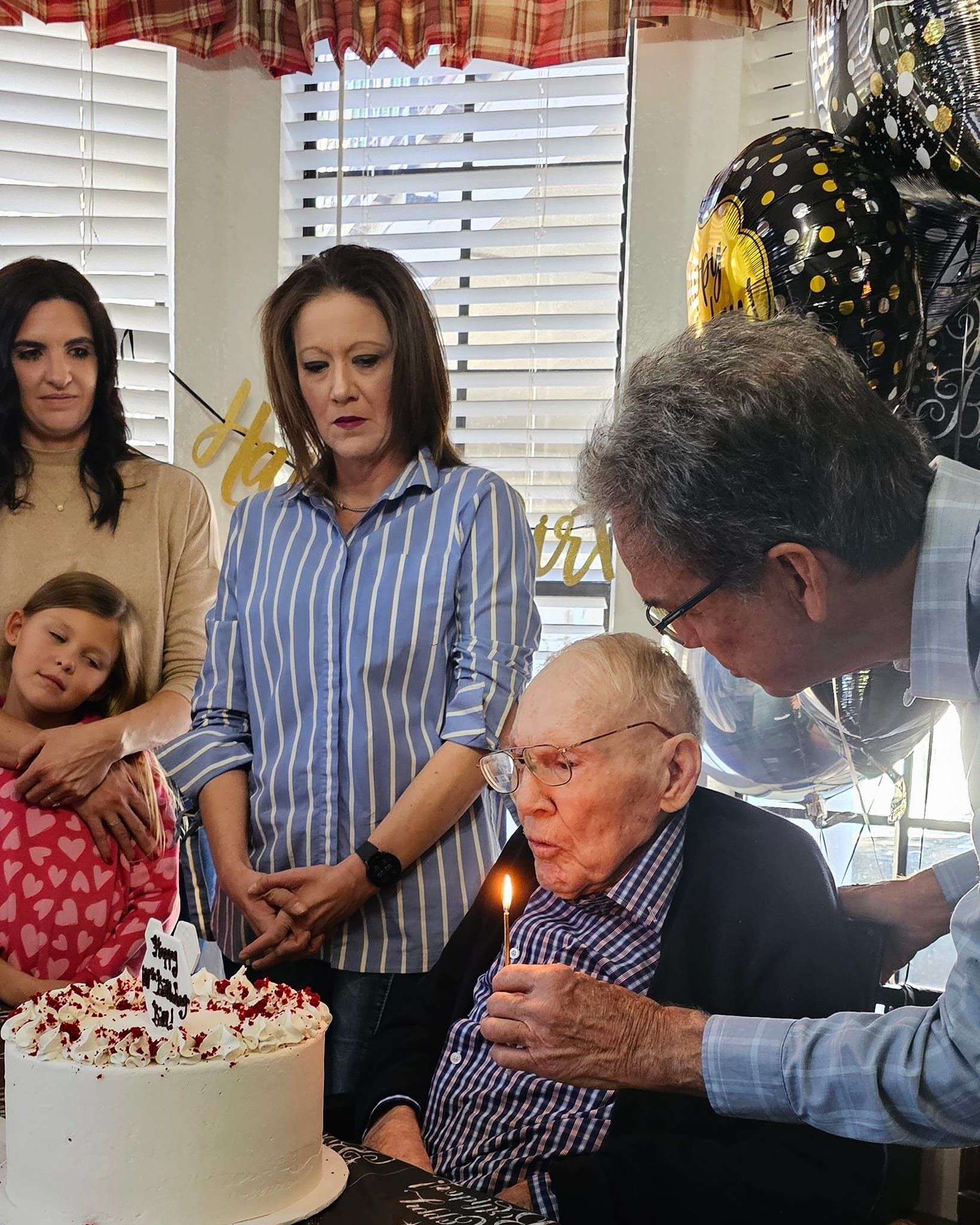 A person lights a candle on a birthday cake for an elderly individual as family watches in a sunlit room.