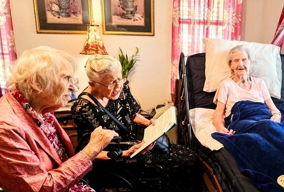 Three people interact in a room, with two sitting and one resting in a hospital bed, one reading from a small book.