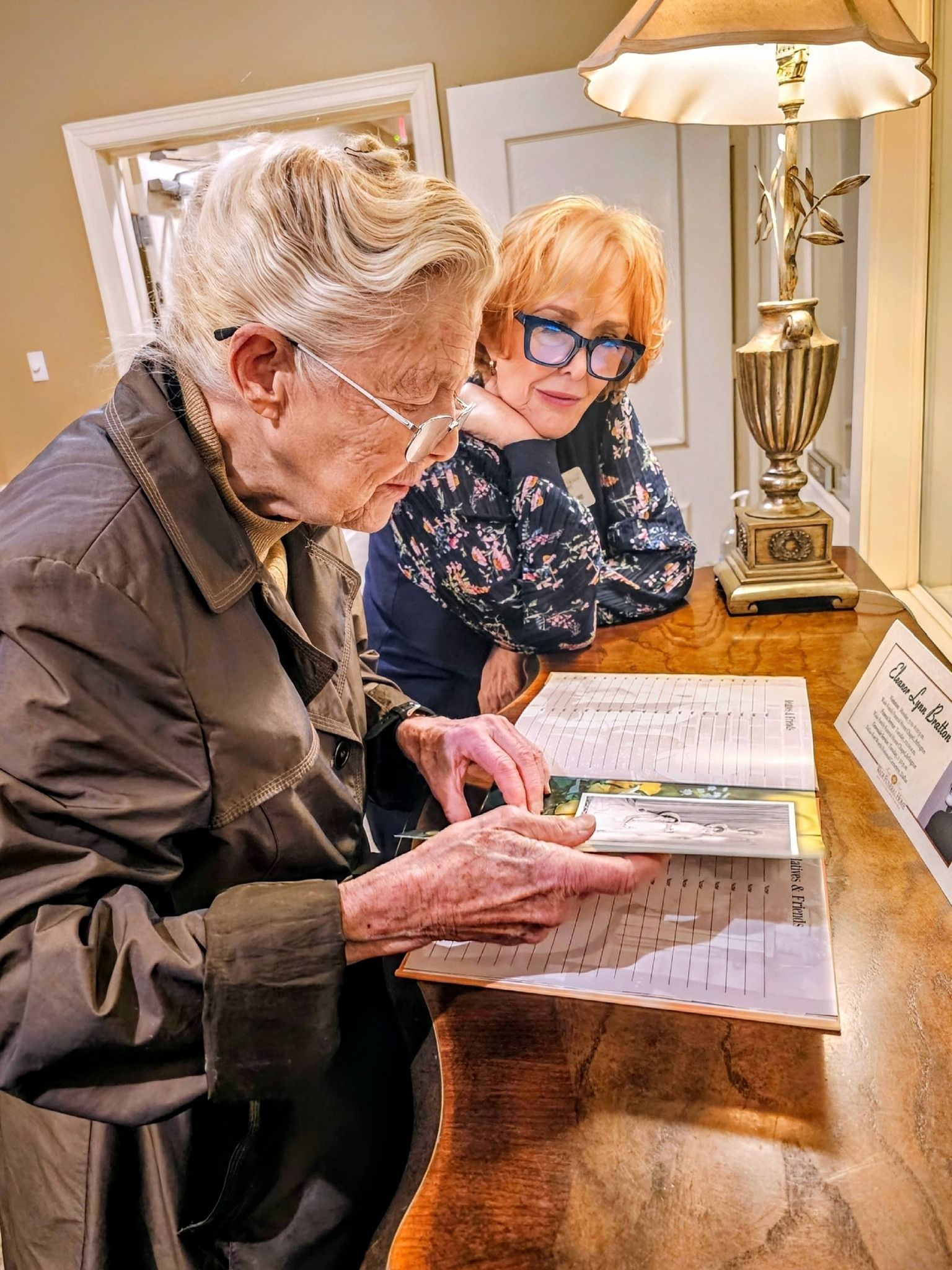 Two individuals looking at a book together on a wooden table under a warm lamp in a softly lit room.