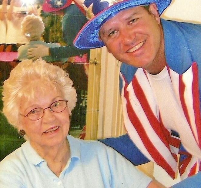 An elderly person sits next to someone wearing a red, white, and blue patriotic hat and jacket, both smiling.