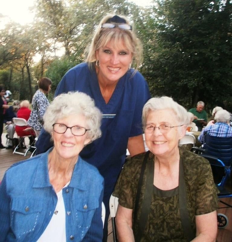 A smiling professional in blue scrubs stands behind two seated people outdoors, both wearing glasses and smiling.